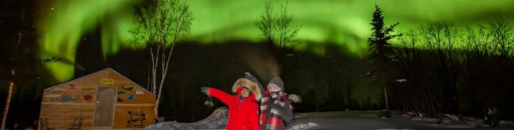 A couple points to the aurora in winter in front of a cabin.